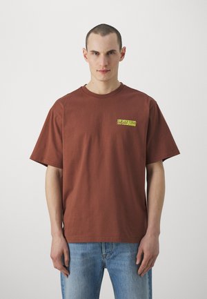 Rjava bombažna majica s kratkimi rokavi in okroglim izrezom, ki ima zeleno grafično tiskanje na prsih. Nosi se z svetlo modrimi kavbojkami.