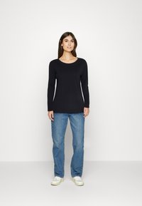 Schwarzes Langarmoberteil mit Rundhalsausschnitt, kombiniert mit hellblauen geraden Jeans und weißen Sneakers, vor einem schlichten Hintergrund.