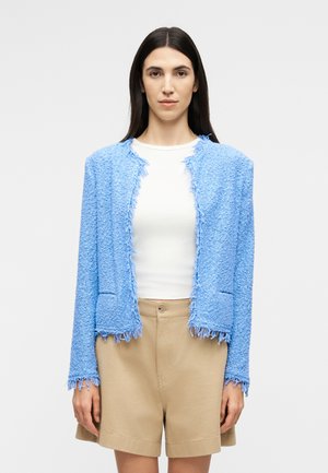 Γυναίκα με μακριά σκουρόχρωμα μαλλιά φοράει ένα υφασμάτινο ανοιχτό μπλε σακάκι, λευκό πουκάμισο και μπεζ σορτς με ψηλή μέση, στέκεται μπροστά σε ένα απλό φόντο.