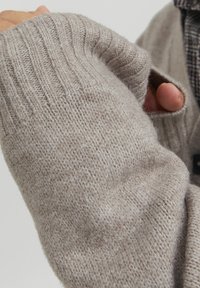 Manica di un maglione lavorato a maglia color beige chiaro, che mette in evidenza la texture a costine e il taglio largo, con un'apertura per il pollice e dettagli di superficie liscia.