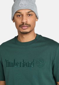 Hombre con gorro gris con logo de árbol blanco y camiseta verde oscuro de Timberland con texto y logo bordados a juego.