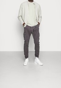 Lichtgrijs overhemd met knopen over een lichtgroen t-shirt, gecombineerd met donkergrijze cargobroeken met zakken en een opgestroopte broekspijp, gedragen met witte sneakers.