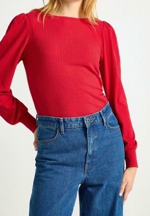 Kobieta ubrana w czerwony, prążkowany top z długim rękawem i bufkami, wsunięty w wysokiej talii niebieskie dżinsy, ręka oparta na biodrze.