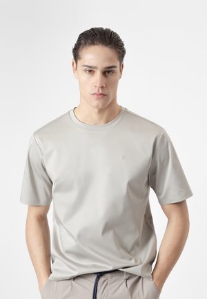 Joven con el cabello peinado hacia atrás, vistiendo una camisa de manga corta gris claro y pantalones beige, de pie con las manos en los bolsillos sobre un fondo blanco.