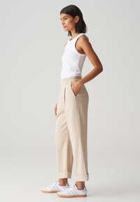 Pantaloni beige sartoriali con vestibilità rilassata, vita elasticizzata, tasche laterali e orli a coste, abbinati a una canotta bianca e sneakers bianche.