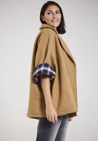 Oversized brown fleece jacket with a collar and plaid cuff accents. Loose fit with a textured surface. Worn over gray jeans.