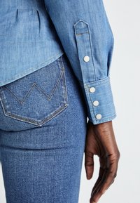 Ljusblå denimskjorta med knäppte ärmar, med synlig sömnad och rundad nederkant. Mörkblå denimjeans med en dekorativ fickdesign.