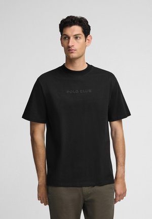 Camiseta negra de algodón con cuello redondo y mangas cortas, que presenta un logo en relieve de "POLO CLUB" en el pecho. Diseño simple y casual.
