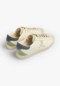 Baskets en cuir crème avec un talon en suède gris et vert. Elles présentent un design à lacets et un logo de skull et os croisés sur le côté.