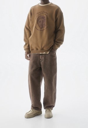 Homme portant un sweat-shirt graphique marron, un pantalon ample marron et des chaussures beige à lacets, debout avec une main dans la poche sur fond blanc.
