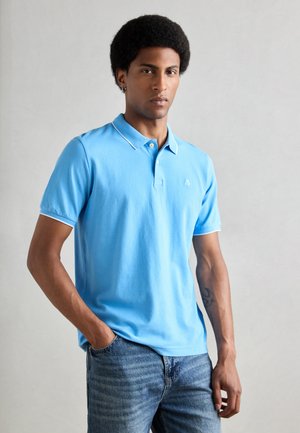 Light blue polo shirt with a collar, short sleeves, and white trim. Emblem on the left chest. Paired with faded blue jeans.