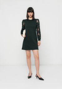 Dark green dress with floral textured sleeves in sheer fabric. Paired with black pointed-toe slingback heels and holding a white clutch.