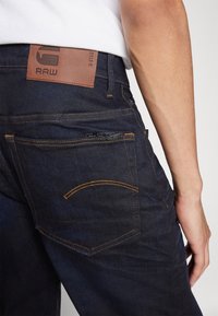 Tmavě modré džíny z denimu s oranžovým prošíváním a koženou značkovou nášivkou na pase, s viditelným detailem zadní kapsy.