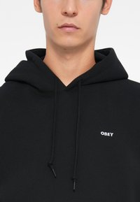 Sort hættejakke lavet af blødt materiale, med en lomme foran, justerbare snore og et lille hvidt "OBEY" logo på brystet.