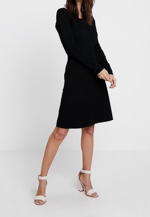Robe noire à manches longues en tissu texturé, présentant une silhouette ajustée et un ourlet à mi-genou. Associée à des talons à bride cheville de couleur claire.