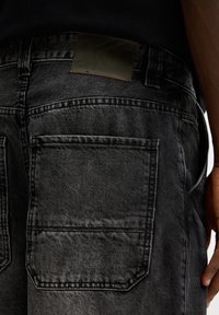Jeans en denim noir avec un tissu texturé présentant des coutures apparentes, un patch en cuir sur la ceinture et deux poches arrière.