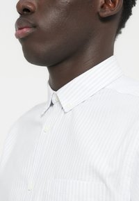 Κοντινό πλάνο του κάτω μέρους του προσώπου και του λαιμού ενός ατόμου που φοράει λευκό πουκάμισο με λεπτές κάθετες ρίγες και γιακά με κουμπιά.