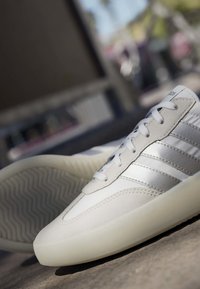 Baskets blanches avec des détails en daim gris et trois bandes argentées reposant sur un sol texturé, inclinées pour montrer le motif de la semelle.
