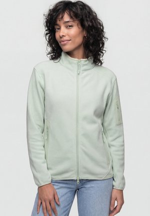Woman with curly hair wearing a light green zip-up fleece jacket and light blue jeans, standing against a plain background.