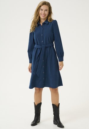 Navy corduroy dress with long sleeves, button front, and a tie belt. Skirt flares slightly below the knee, paired with black cowboy boots.