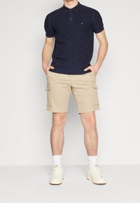 Marineblauw poloshirt met korte mouwen en een gestructureerd patroon, gecombineerd met beige cargoshorts met zijzakken. Gedragen met witte sneakers en sokken.
