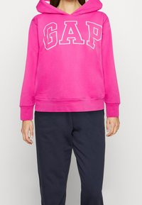 Sudadera con capucha rosa con letras blancas "GAP" en el pecho. Presenta puños y dobladillo acanalados, hecha de una tela suave de peso medio.