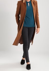 Brązowy wełniany płaszcz na tealowej bluzce, zestawiony z czarnymi obcisłymi spodniami i czarnymi butami wsuwanymi. Płaszcz ma długi krój i zapięcie na guziki.