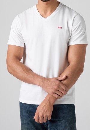 Mężczyzna ubrany w białą koszulkę V-neck marki Levi’s i niebieskie dżinsy, trzymający lewe nadgarstek prawą ręką na jednolitym szarym tle.