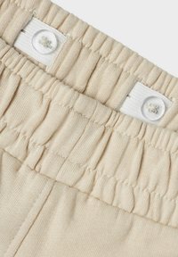 Ceinture en tissu beige avec une texture froncée, dotée d'un bouton blanc et d'un élastique pour ajustement. Convient pour un port confortable.