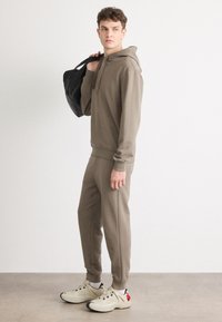 Hanorac bej cu buzunar de tip kangaroo, pantaloni jogger tapered asortați și adidași crem cu accente negre și roșii. Modelul ține o geantă tote neagră.