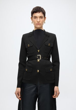 Kvinde iført en tætsiddende sort blazer med guldknapper og bælte, kombineret med sorte bukser og guldringeøreringe, foran en ensfarvet baggrund.