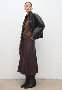 Veste en cuir noire avec détails en boutons, pull en maille marron et jupe longue bordeaux. Associé avec des bottes noires à la cheville. Textures lisses.