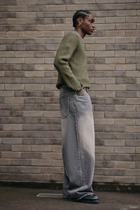 Olivgrön tröja med texturerad stickning, kombinerad med vida grå jeans med en bakficka. Svarta klumpiga skor fulländar looken.