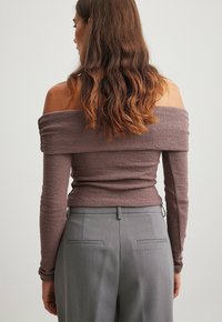 Kvinna med vågigt brunt hår som bär en mauve off-shoulder långärmad topp och högmidjade grå byxor, vänd bort från kameran mot en enkel bakgrund.