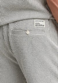 Stribede stofshorts med et sort-hvidt lodret stribemønster, knaplukning på lommen og et mærkat med tekstbeskrivelse.
