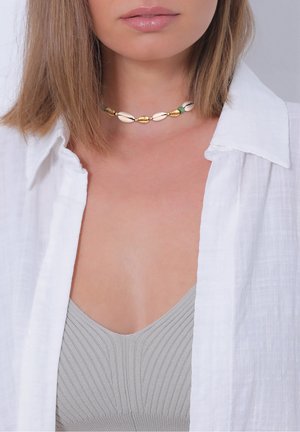 Femme portant une chemise blanche ouverte, un haut gris côtelé et un collier ras du cou composé de perles de coquillage alternant blanc et or, avec une perle verte.