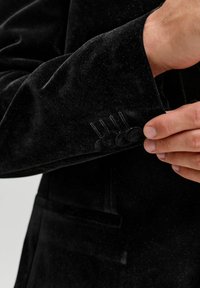 Juodo aksominio švarko rankovė su trimis mygtukais rankogalyje, subtiliais siuvimo detalėmis ir lygiu tekstūra.