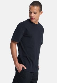 Jeune homme en t-shirt noir et pantalon, regardant vers le bas et sur le côté, les mains dans les poches, devant un fond blanc uni.