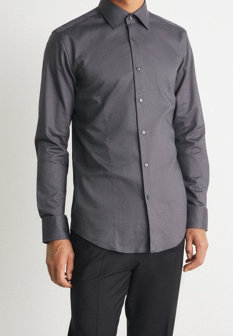 Homme portant une chemise ajustée à manches longues gris foncé avec col et pantalon noir, debout devant un fond clair uni.