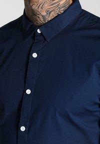 Uomo che indossa una camicia blu navy con bottoni bianchi e un tatuaggio visibile sul collo e sulla parte superiore del torace.