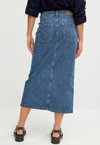 Gonna di jeans di colore blu scuro, di lunghezza media, con tasche posteriori, un taglio dritto e dettagli di cucitura sottile. Indossata con sandali con zeppa neri.