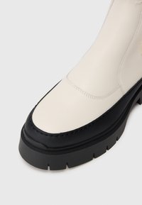 Botine din piele sintetică albă, cu o talpă din cauciuc negru, prezentând cusături contrastante și un șablon texturat pentru aderență.