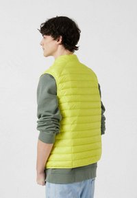Lichte gewatteerde bodywarmer in fel limegroen, met horizontale stiksels, een hoge kraag, gedragen over een grijze sweatshirt met lange mouwen.