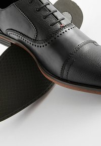 Zapato de vestido de cuero negro con acentos perforados, costuras detalladas y una suela de goma plana con un patrón texturizado.