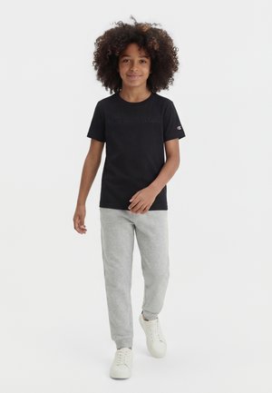 Child with curly hair wearing a black Champion T-shirt, grey sweatpants, and white sneakers, standing and smiling on a white background.