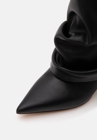 Botte en cuir noire à bout pointu et effet décontracté avec des plis doux autour de la cheville, présentée sur fond blanc.