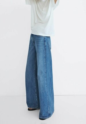 Osoba ubrana w szerokie niebieskie dżinsy z denimu i luźną białą koszulę, stojąca na tle jednolitego białego tła.