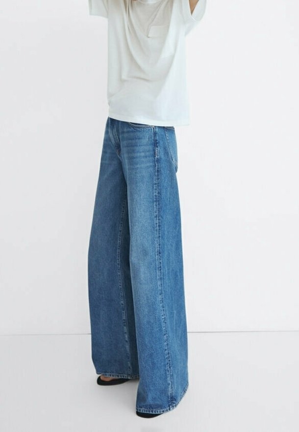 Personne portant un jean bleu à jambes larges et une chemise blanche ample, debout devant un fond blanc uni.