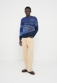 Ναυτικό πλεκτό πουλόβερ με μπλε διαβάθμιση, γραφικό σχέδιο αγωνιστικού αυτοκινήτου, ριμπ μανσέτες και τελείωμα· σετ με φαρδιά παντελόνια ανοιχτού μπεζ χρώματος και καφέ παπούτσια.