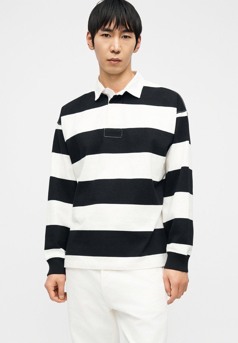 Jeune homme portant un polo à manches longues à larges rayures noires et blanches avec un col blanc et un pantalon blanc, debout devant un fond uni.